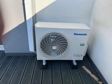Zewnętrzna jednostka klimatyzacji Panasonic zamontowana na czarnych podkładkach na drewnianym tarasie, widoczny fragment ściany budynku i oznaczenie czynnika chłodniczego R32.