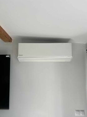 Biała jednostka klimatyzacji Panasonic zawieszona na szarej ścianie w jasnym wnętrzu, widoczny fragment drewnianej belki stropowej i czarny telewizor z lewej strony.