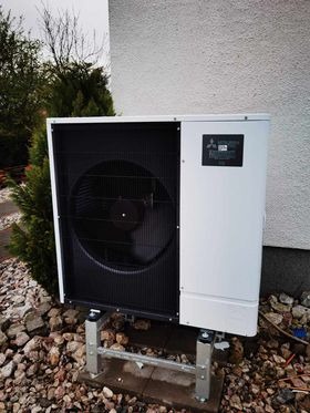 Zewnętrzna jednostka pompy ciepła Mitsubishi Ecodan na metalowych wspornikach, otoczona kamieniami i zielenią, przy białej ścianie budynku.