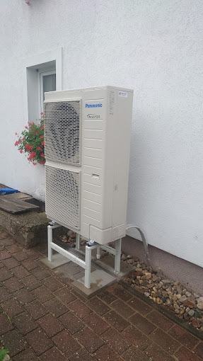 Zewnętrzna jednostka pompy ciepła Panasonic Inverter zamontowana na metalowej konstrukcji na ceglanym chodniku, obok ściany budynku z oknem i kwiatami.