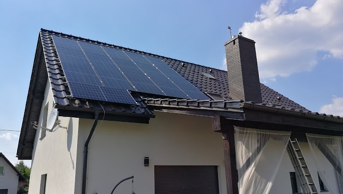 Panele fotowoltaiczne na dachu domu jednorodzinnego z ciemną dachówką, widoczne anteny satelitarne i drabina oparta o ścianę.