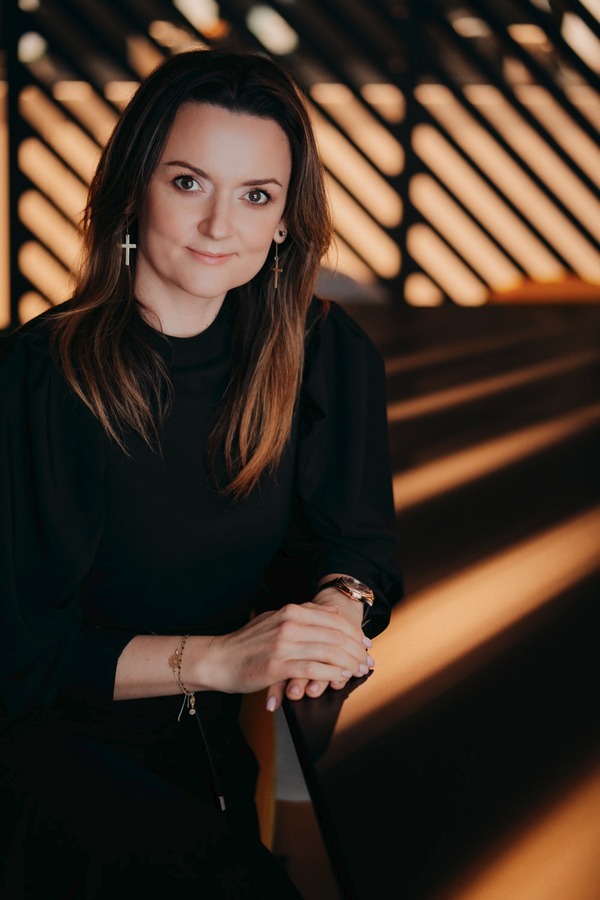 Kobieta w czarnej bluzce, z delikatnym uśmiechem, pozuje na portrecie biznesowym w otoczeniu geometrycznych cieni, nosząc złote kolczyki w kształcie krzyży i bransoletkę.