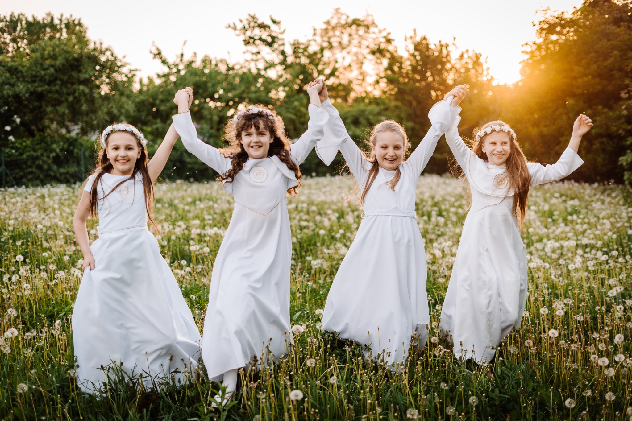 Cztery dziewczynki w białych strojach komunijnych, trzymające się za ręce, radośnie biegają po łące pełnej dmuchawców w promieniach zachodzącego słońca.