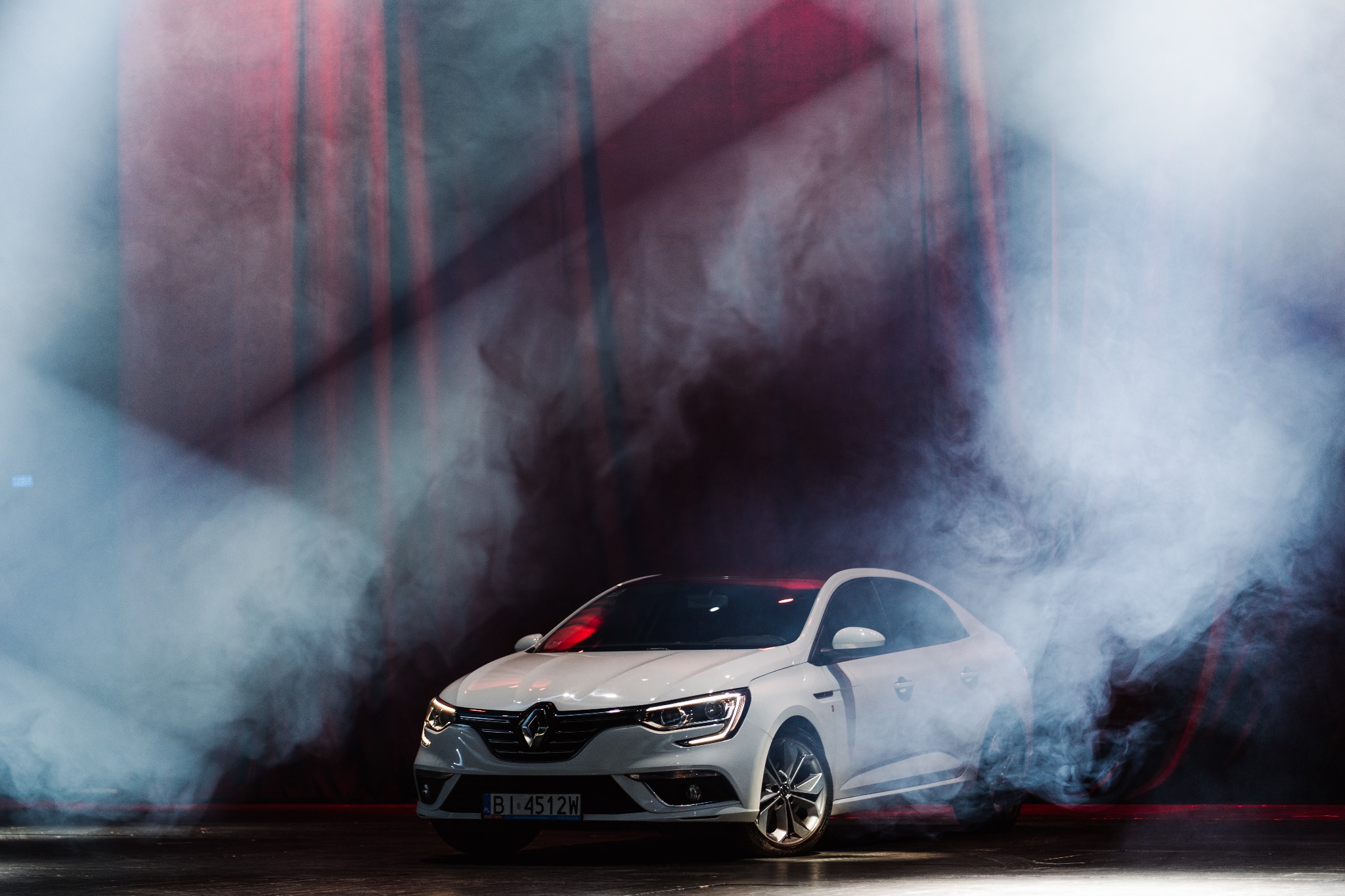 Biały samochód marki Renault w studiu, otoczony dymem, z widocznymi reflektorami i czerwonymi akcentami świetlnymi w tle.