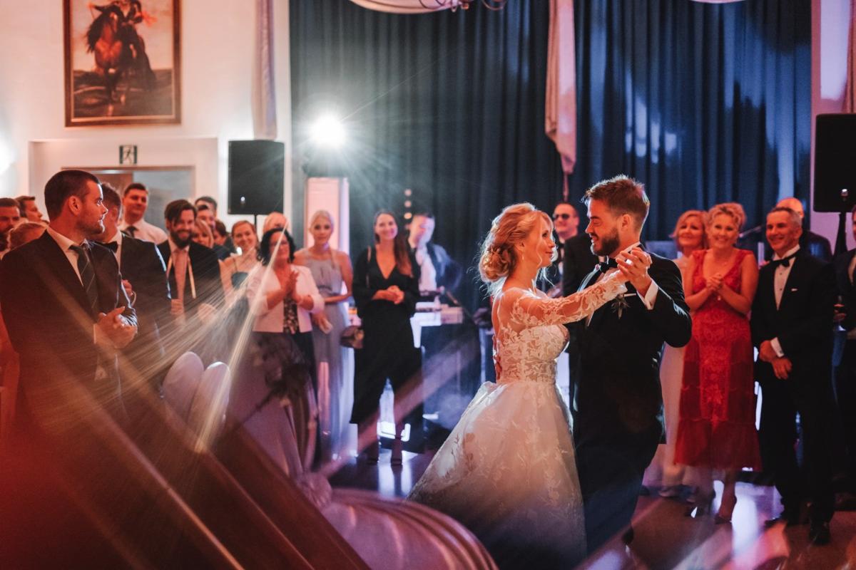 Para młoda tańczy pierwszy taniec na weselu, w tle goście i oświetlenie sceniczne.