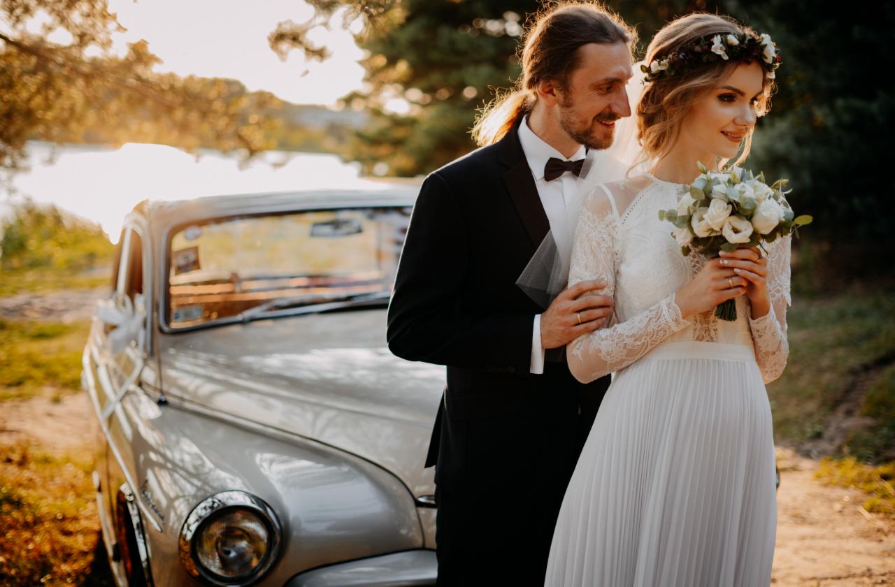 Para młoda w strojach ślubnych pozuje przy zabytkowym, jasnym samochodzie na tle jeziora i drzew w złocistym świetle.