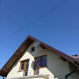 Antena satelitarna zamontowana na ścianie domu z brązowym dachem i drewnianymi oknami, widok od dołu w słoneczny dzień.