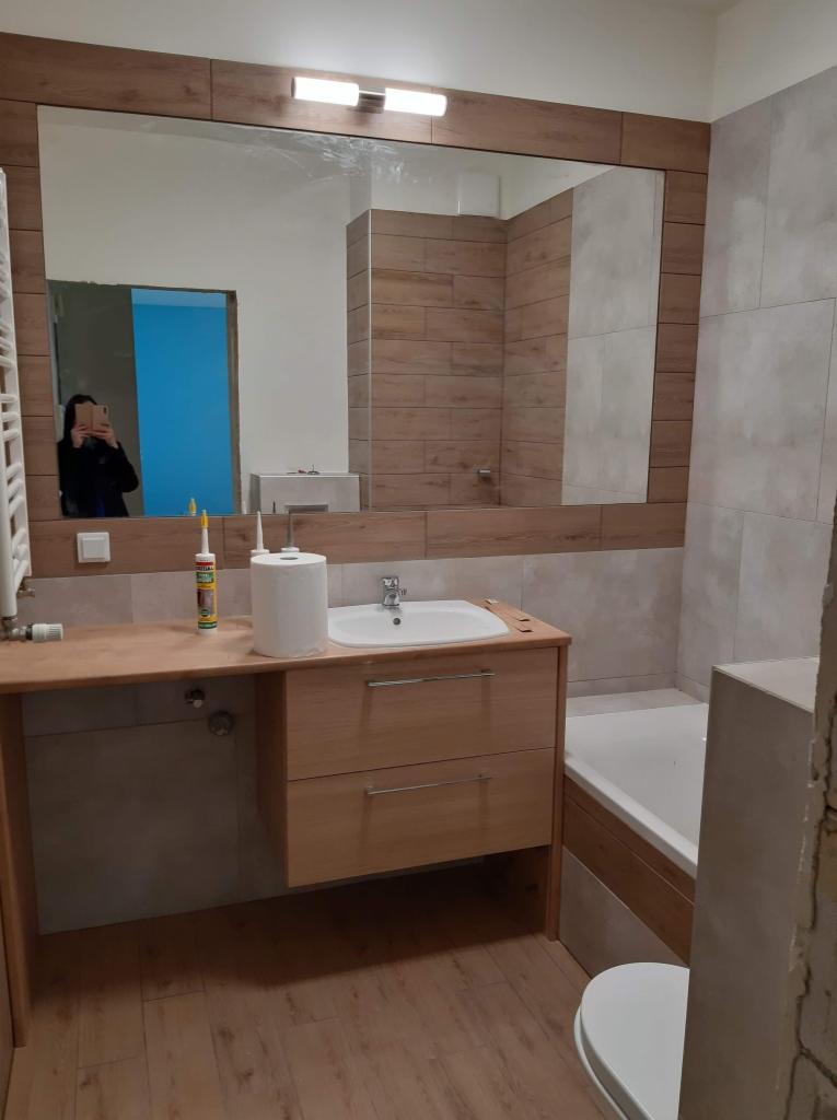 Wykończona łazienka z drewnopodobnymi płytkami na ścianach, drewnianą szafką pod umywalką i dużym lustrem, w którym odbija się osoba robiąca zdjęcie w innym, niebieskim pomieszczeniu.