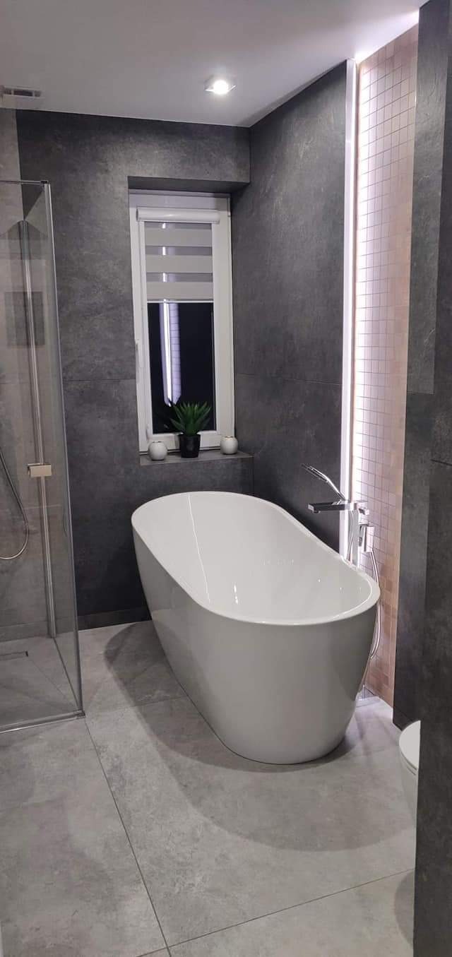 Nowoczesna łazienka w Piotrkowie Trybunalskim z wolnostojącą wanną, szarą glazurą imitującą kamień, kabiną prysznicową ze szklanymi drzwiami i oknem z roletami w tle.