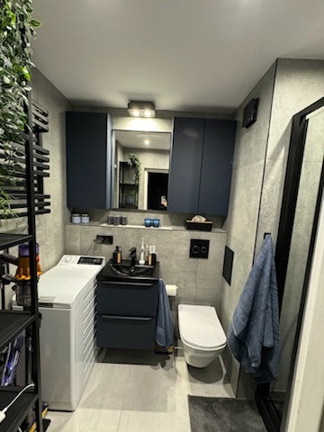 Nowoczesna łazienka z szarymi płytkami, ciemnoniebieskimi szafkami i czarną armaturą, pralka obok umywalki, dekoracyjne rośliny w rogu.