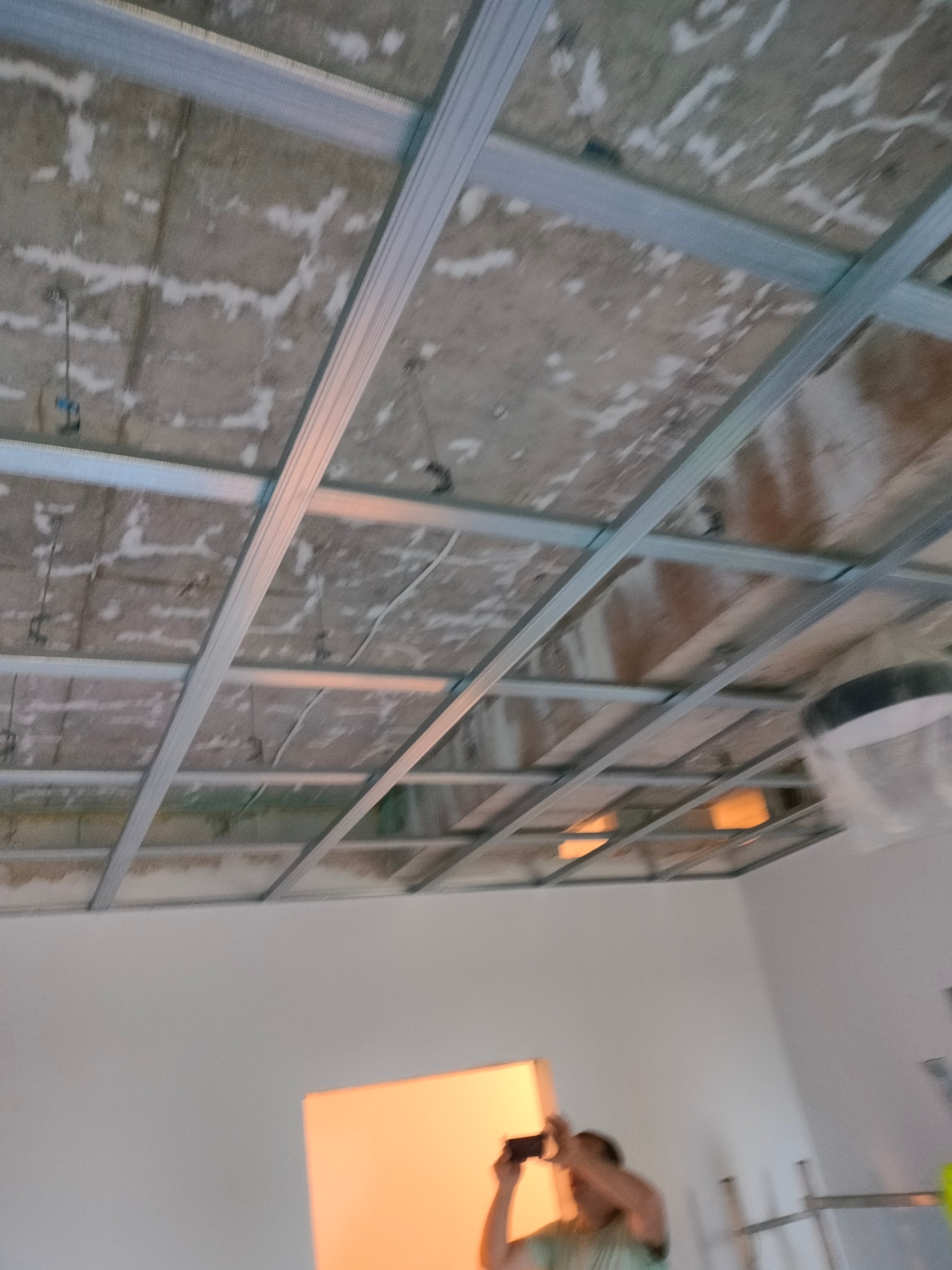 Stelaż sufitowy z profili metalowych, widoczny surowy strop i fragment ściany z oświetleniem, osoba robiąca zdjęcie w tle.