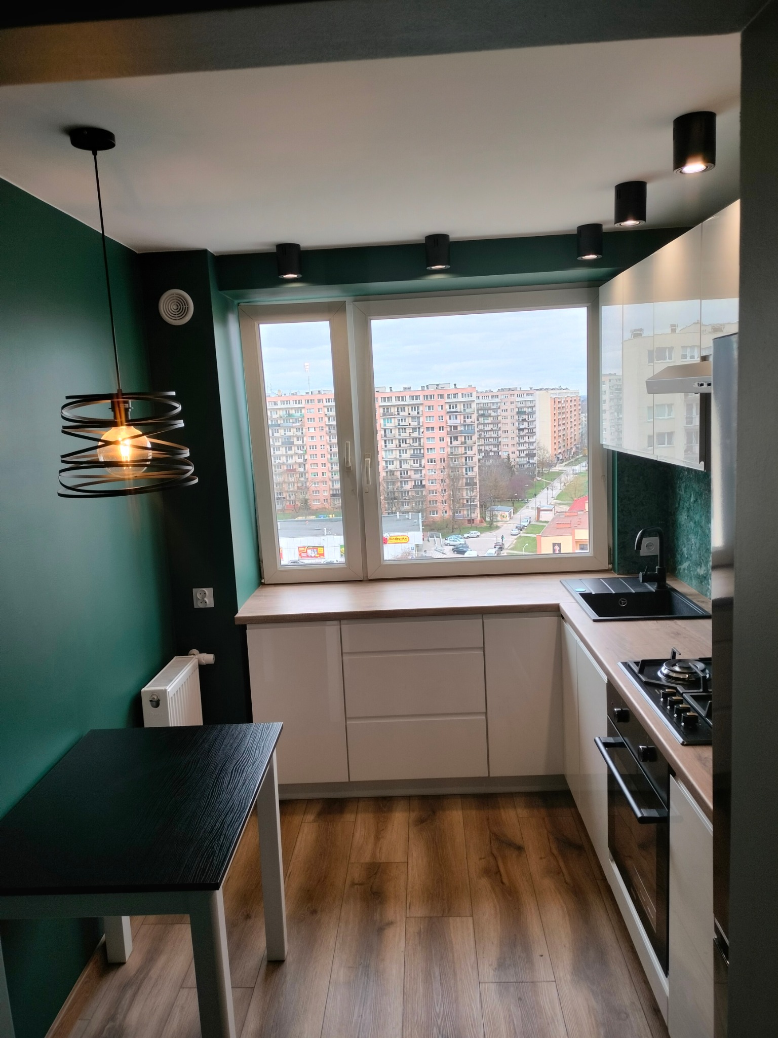 Nowoczesna kuchnia z białymi szafkami, drewnianym blatem i zieloną ścianą, widok na miasto przez duże okno.