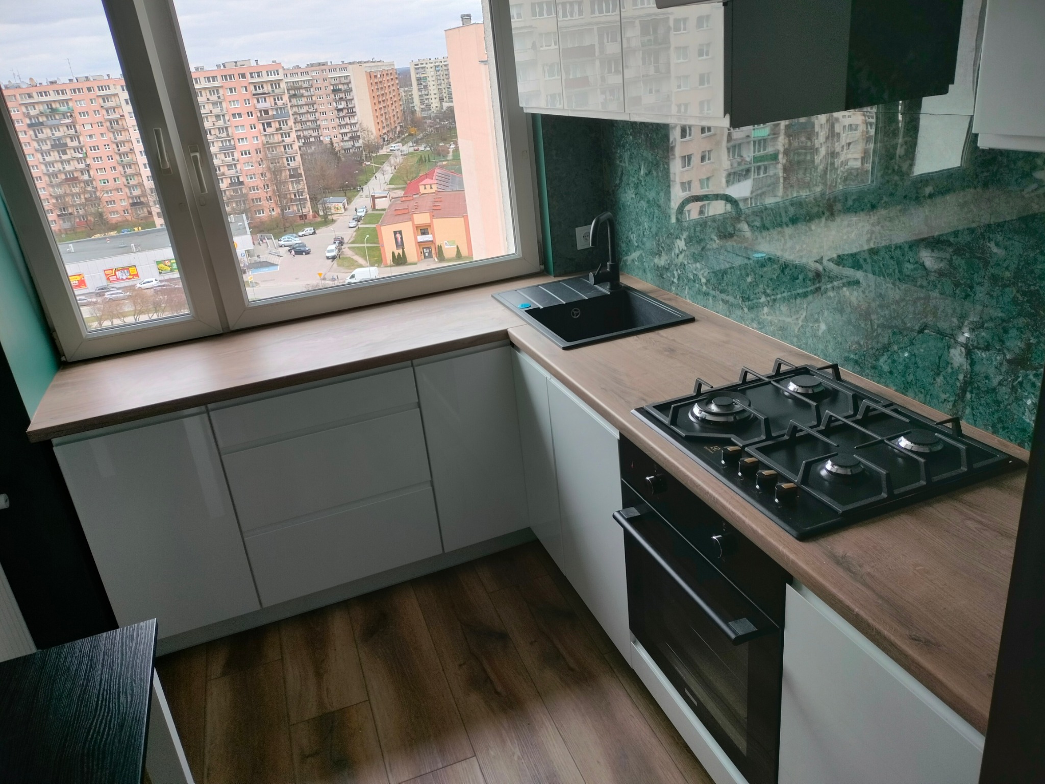 Nowoczesna kuchnia z białymi szafkami, drewnianym blatem i zielonym marmurowym backsplashiem, widok na miasto przez okno.