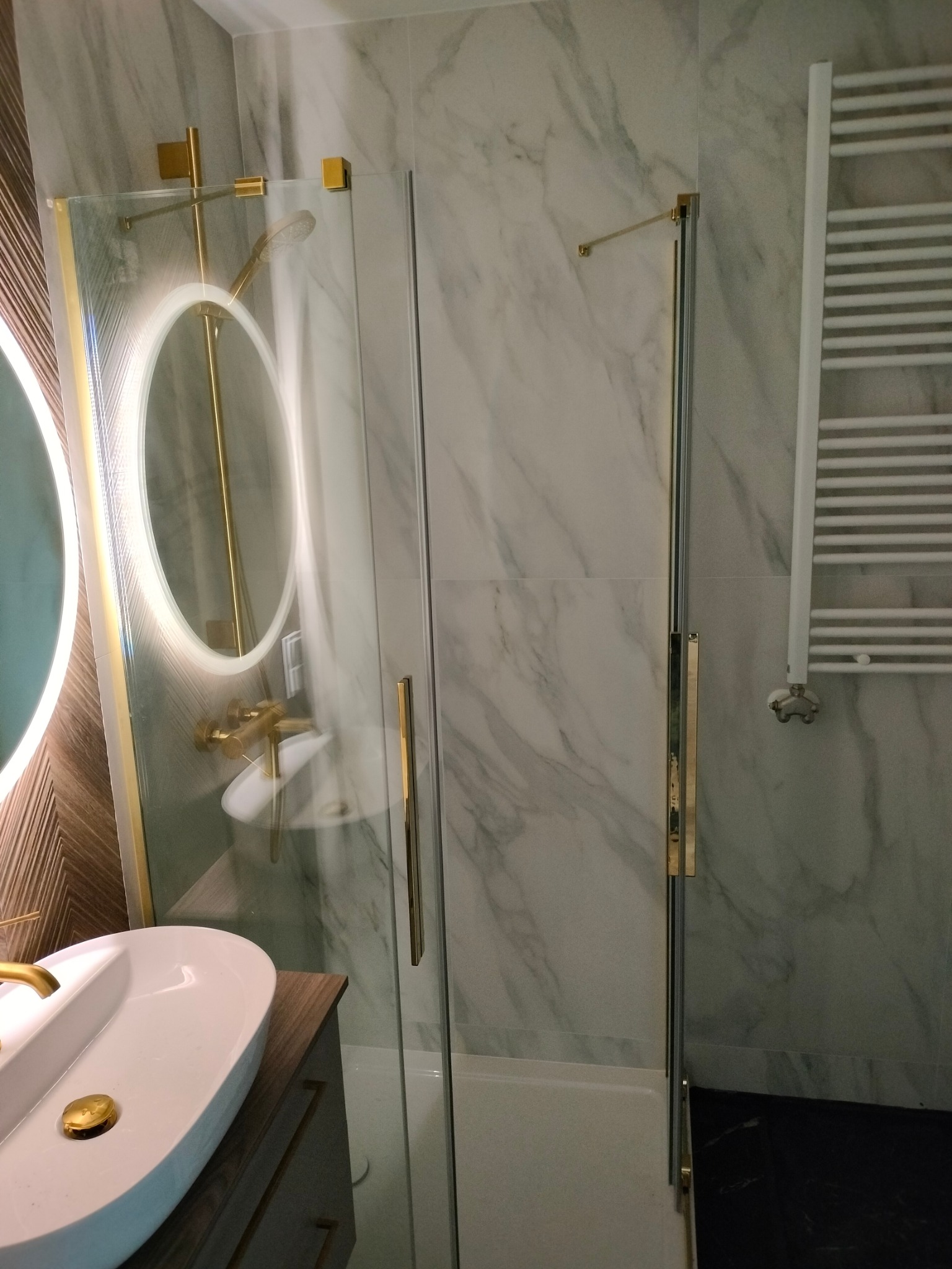 Nowoczesna kabina prysznicowa ze złotymi detalami, umywalka z baterią w podobnym stylu, marmurowe płytki na ścianach i grzejnik drabinkowy w wyremontowanej łazience.