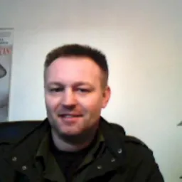 Portret mężczyzny w ciemnej kurtce na tle jasnej ściany i plakatu, uśmiechający się do kamery.