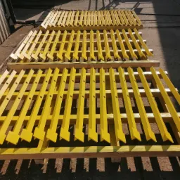 Żółte, metalowe elementy konstrukcyjne z otworami, ułożone na drewnianych paletach w rzędach, prawdopodobnie po malowaniu.