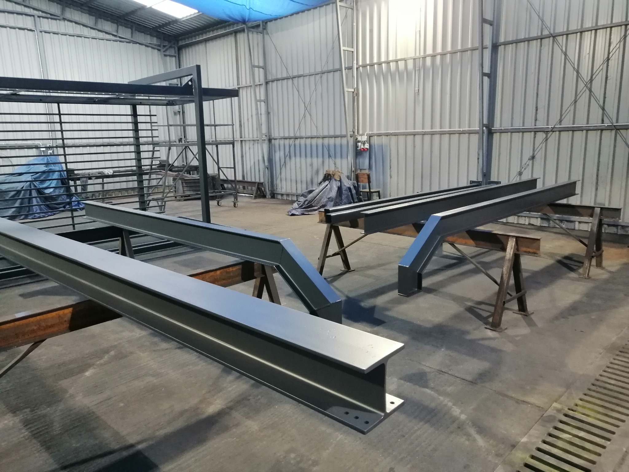 Metalowe elementy konstrukcyjne, prawdopodobnie elementy szkieletu hali, leżące na kozłach w hali produkcyjnej; widoczne spawane łączenia i otwory montażowe.
