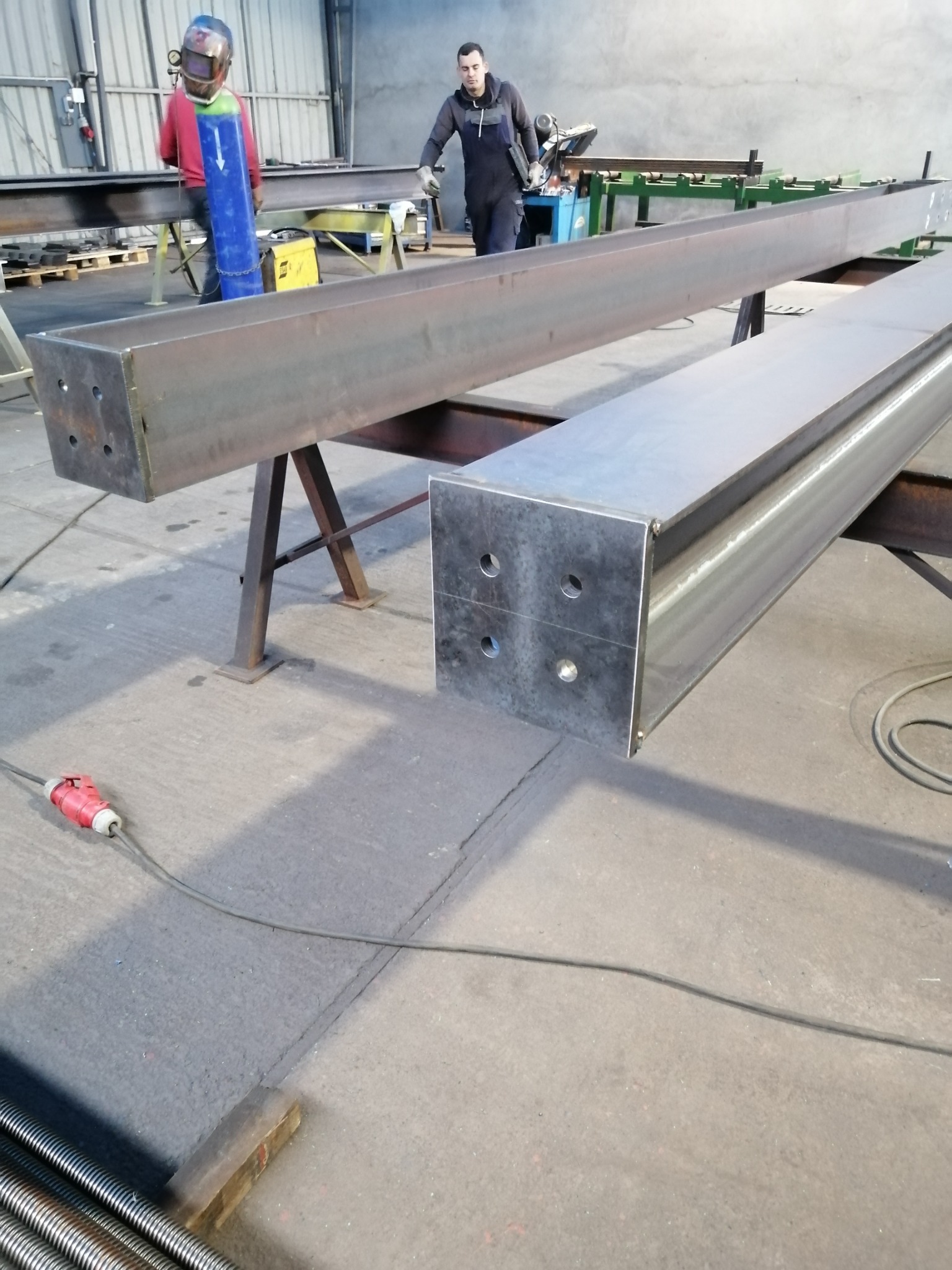 Dwie długie, stalowe belki o przekroju prostokątnym na metalowych kozłach w hali, spawacz w masce i kombinezonie w tle, widoczne otwory montażowe w końcach belek.