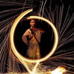 Mężczyzna w okularach i kimonie, otoczony wirującymi iskrami ognia, tworzącymi świetlisty okrąg, podczas pokazu pirotechnicznego.