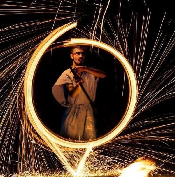 Mężczyzna w okularach i kimonie, otoczony wirującymi iskrami ognia, tworzącymi świetlisty okrąg, podczas pokazu pirotechnicznego.