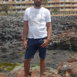 Mężczyzna w okularach przeciwsłonecznych stoi na skałach w klapkach, w tle budynek z balkonami i palmami. Ubrany w koszulkę z nadrukiem i krótkie spodenki.