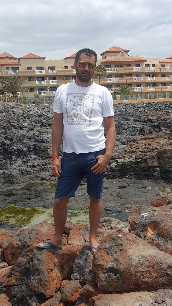 Mężczyzna w okularach przeciwsłonecznych stoi na skałach w klapkach, w tle budynek z balkonami i palmami. Ubrany w koszulkę z nadrukiem i krótkie spodenki.