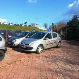 Srebrny samochód Renault Clio z oznaczeniem 'L' na dachu i logo 'Turbo' na boku, zaparkowany na czerwonej kostce, w tle inne auta i zieleń.
