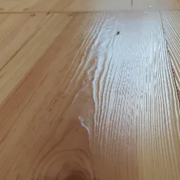 Jasna, drewniana podłoga z wyraźnym usłojeniem i widocznym sękiem, oświetlona naturalnym światłem wpadającym z góry.