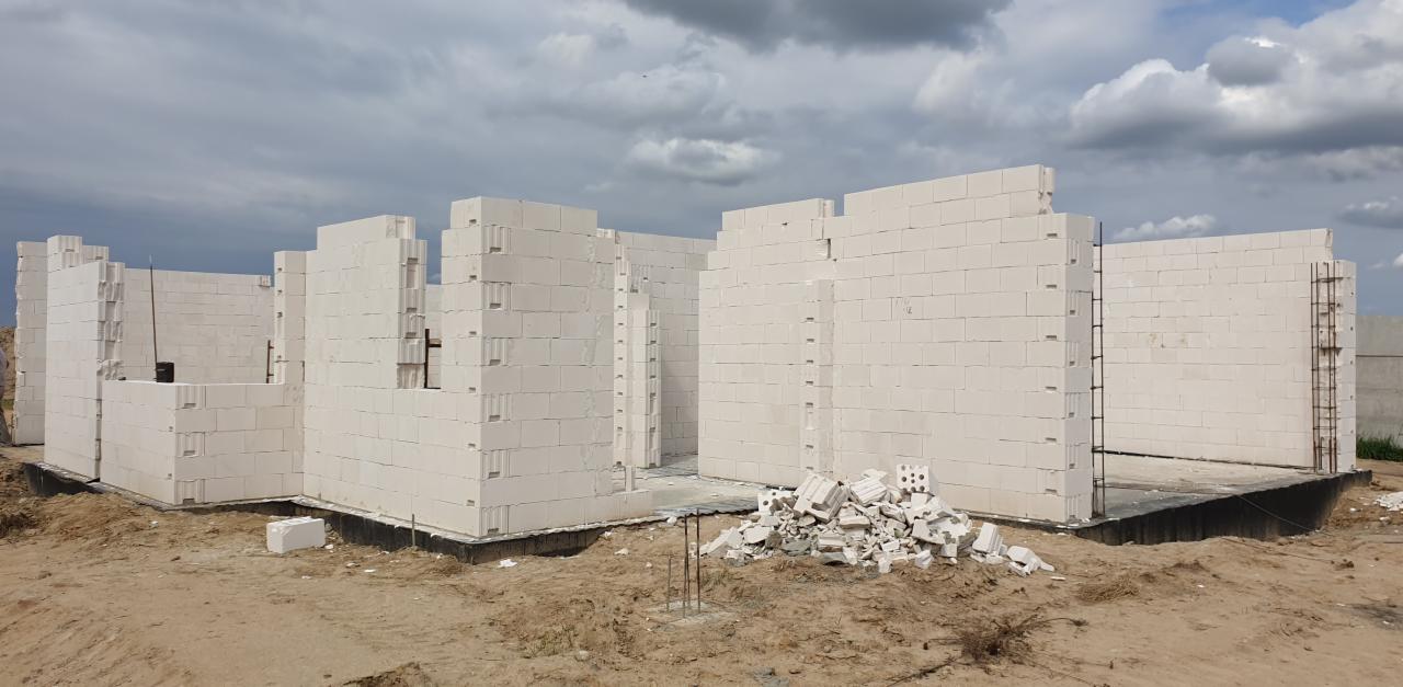 Wznoszone ściany parteru domu z białych bloczków betonu komórkowego, widoczne zbrojenia pionowe, na pierwszym planie sterta gruzu z tych samych bloczków, pochmurne niebo.