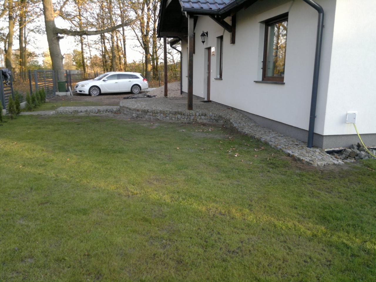 Ułożona kostka brukowa wokół domu z białymi ścianami i samochodem w tle, widok z trawnika.