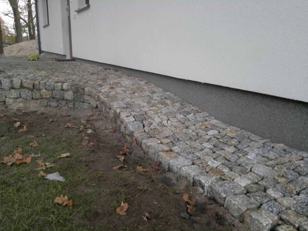 Ścieżka z kostki granitowej ułożona wzdłuż białej ściany budynku, z widocznymi opadłymi liśćmi na trawie i ziemi.