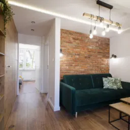 Salon z ceglaną ścianą, zieloną sofą i drewnianą podłogą, widok na przedpokój i pokój z oknem, oświetlenie LED i wiszące żarówki.