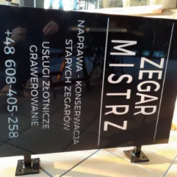 Czarna tablica reklamowa z białym tekstem 'ZEGAR MISTRZ', 'NAPRAWA - KONSERWACJA STARYCH ZEGARÓW', 'USŁUGI ZŁOTNICZE GRAWEROWANIE' oraz numerem telefonu, umieszczona na metalowych nóżkach.
