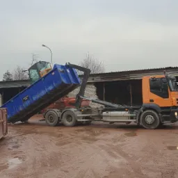 Pomarańczowa ciężar&oacute;wka z niebieskim kontenerem przechylonym do g&oacute;ry, stojąca na błotnistym terenie, z widocznymi śladami opon