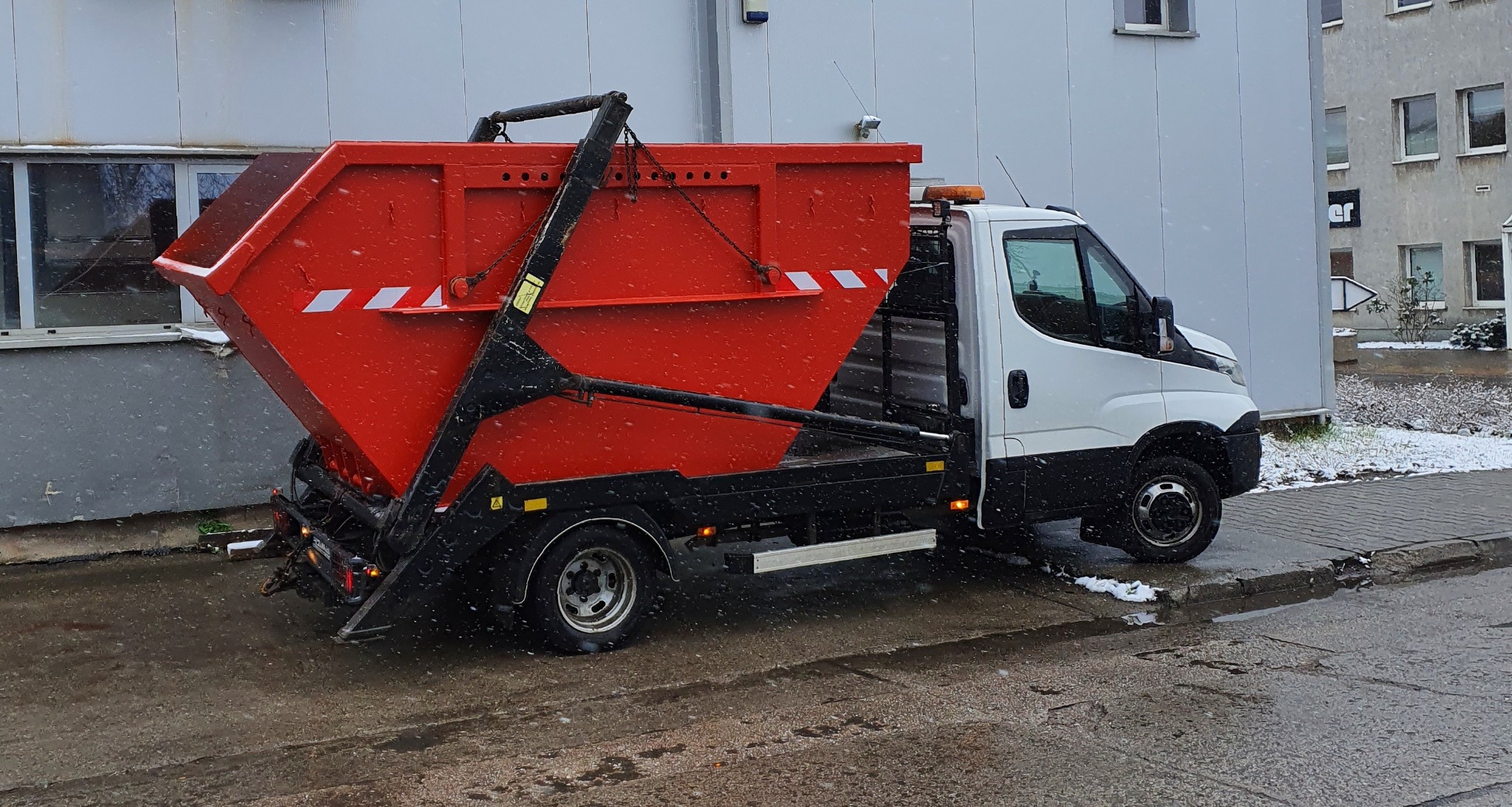 Ciężarówka z czerwonym kontenerem na platformie podczas lekkiego opadu śniegu, zaparkowana przy szarym budynku.