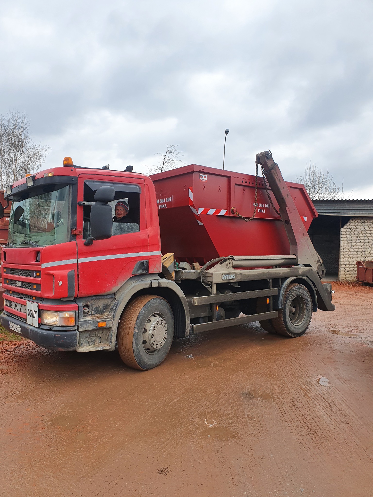 Czerwony samochód ciężarowy Scania z kontenerem na gruz, kierowca w kabinie, błotniste podłoże.