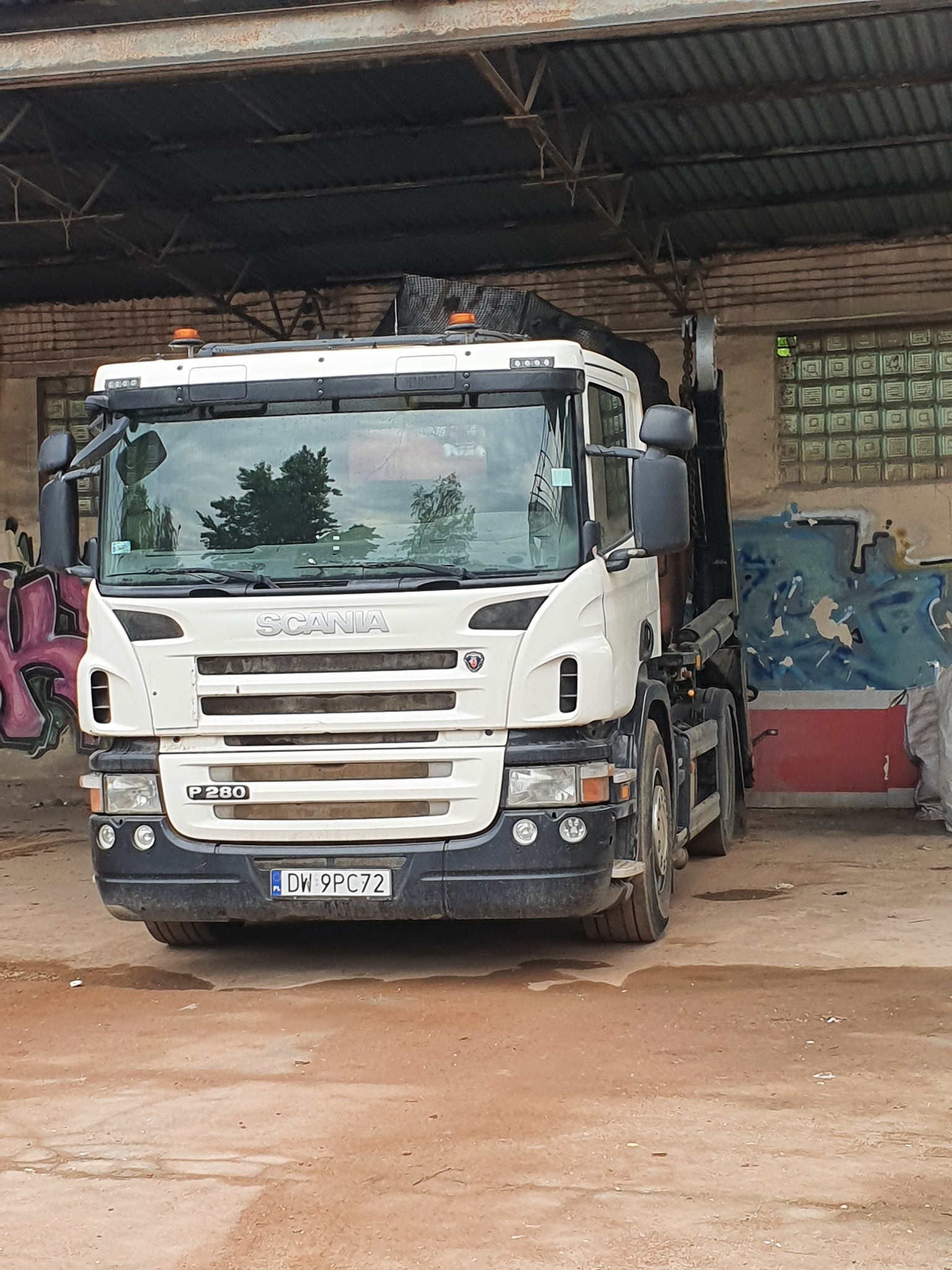 Biała ciężarówka Scania P280 z podnośnikiem hakowym, zaparkowana w zadaszonym, częściowo zrujnowanym magazynie, z widocznymi graffiti na ścianach i ziemią pokrytą plamami.