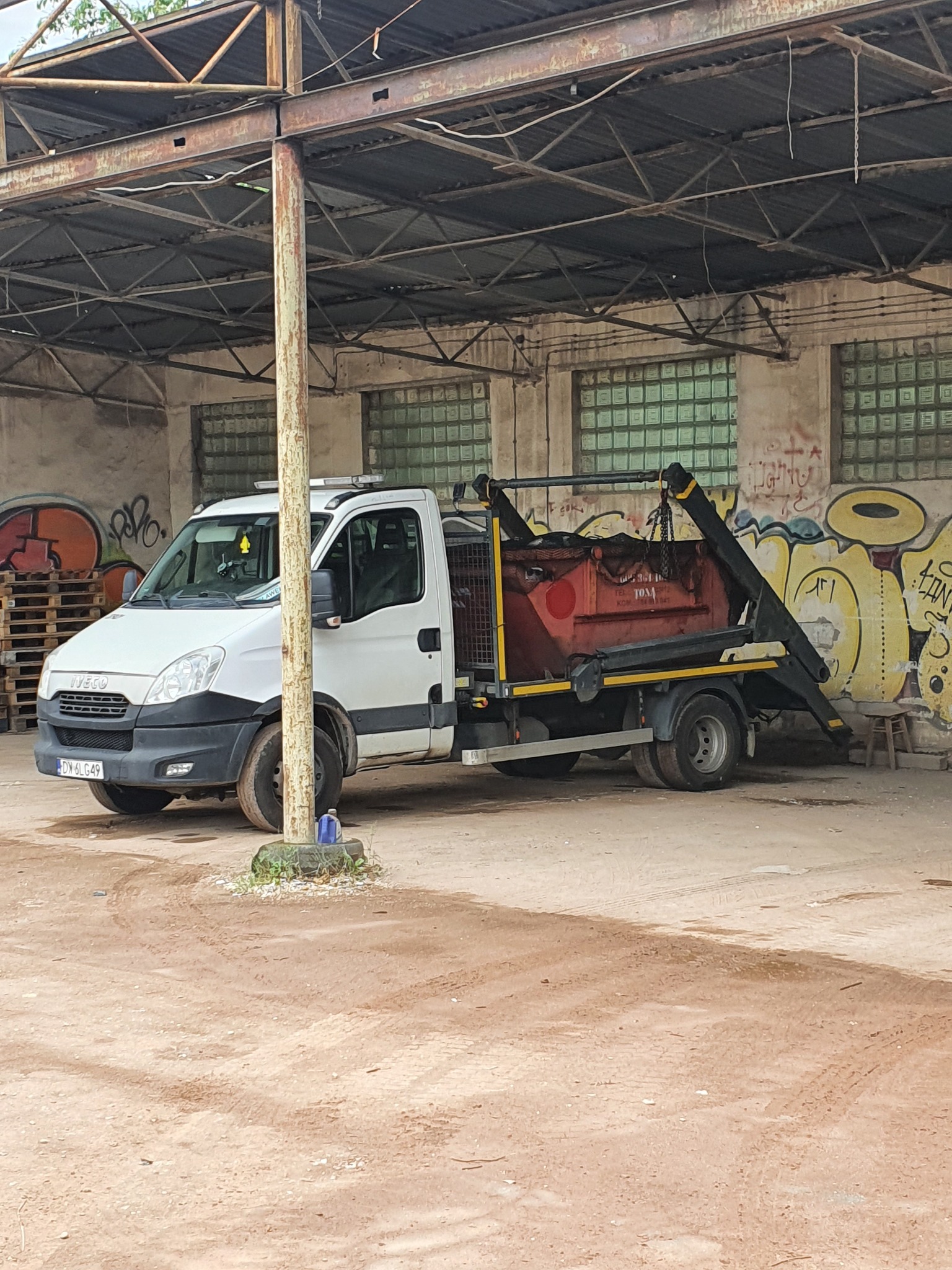 Biały samochód ciężarowy z kontenerem na hakowcu stoi na placu pod zadaszeniem, w tle ściana z graffiti.