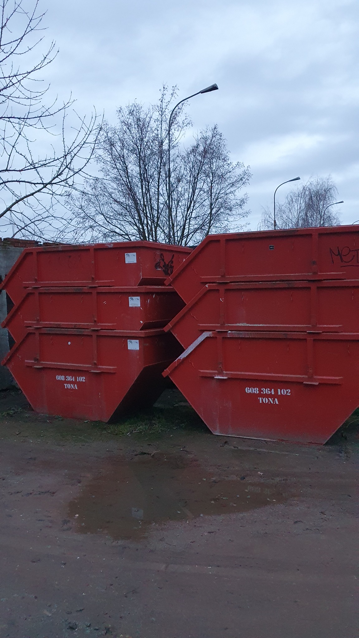 Stos trzech czerwonych kontenerów z numerem telefonu i napisem 'TONA', ustawionych na ziemi z kałużą, na tle szarego nieba i drzew bez liści.