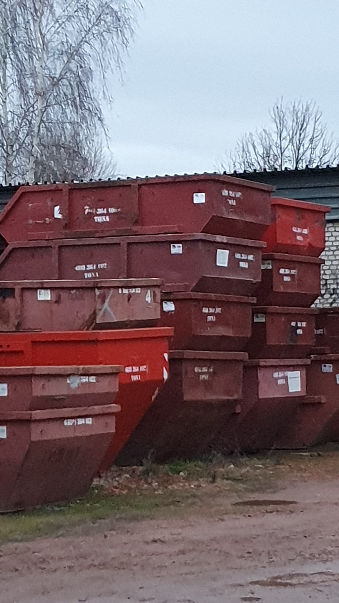 Stos czerwonych, pustych kontenerów ustawionych jeden na drugim na ziemi, z widocznymi napisami i numerami identyfikacyjnymi, na tle szarego nieba i drzew bez liści.