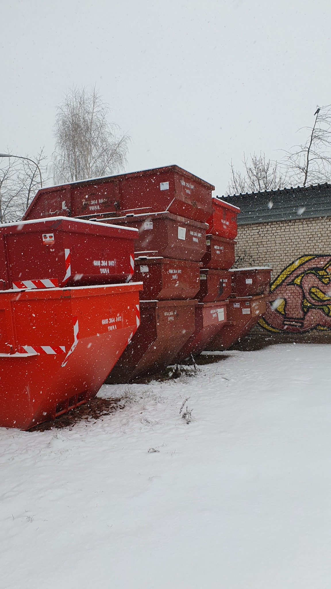 Stos czerwonych kontenerów na gruz ustawionych na śniegu podczas opadów, widoczny numer telefonu i biało-czerwone pasy na jednym z kontenerów, w tle mur z graffiti.