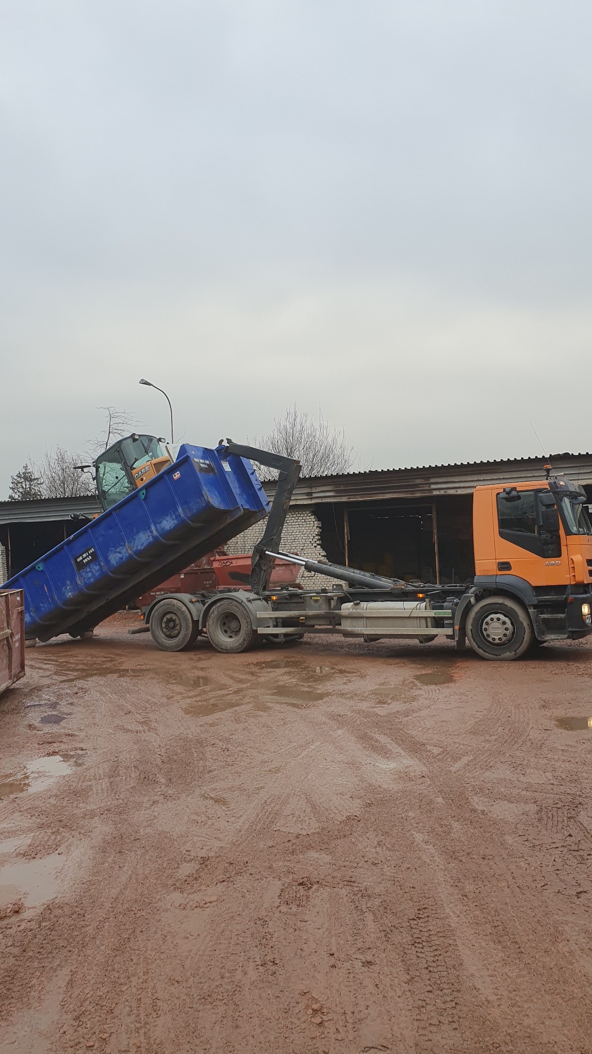 Pomarańczowa ciężarówka z niebieskim kontenerem przechylonym do góry, stojąca na błotnistym terenie, z widocznymi śladami opon