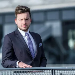 Portret mężczyzny w garniturze, opierającego się o metalową barierkę na tle nowoczesnego budynku.