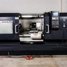 Czarna tokarka CNC marki Janus z panelem sterowania i czerwonym pedałem nożnym, oświetlona jarzeniówką, ustawiona na betonowej podłodze w hali.