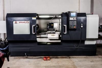 Czarna tokarka CNC marki Janus z panelem sterowania i czerwonym pedałem nożnym, oświetlona jarzeniówką, ustawiona na betonowej podłodze w hali.