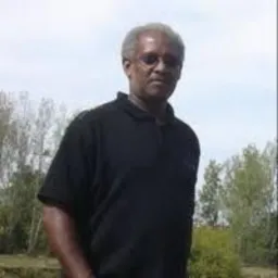 Portret mężczyzny w czarnej koszulce polo na tle drzew w Polska Cerekiew. Mężczyzna ma siwe włosy i okulary przeciwsłoneczne. Ujęcie od pasa w górę.