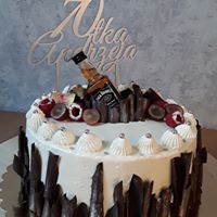 Tort urodzinowy z białym kremem, dekorowany czekoladowymi wiórkami, owocami i miniaturową butelką Jack Daniel's, z topperem z imieniem na 70 urodziny.