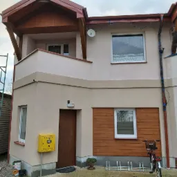 Dwukondygnacyjny dom bliźniak po tynkowaniu elewacji w odcieniach beżu, z drewnianą okładziną, widoczne rusztowanie i zabezpieczone rynny.