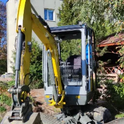 Żółto-szara mini koparka na trawniku przed budynkiem, częściowo zagłębiona w ziemi, sugerująca prace ziemne w otoczeniu zieleni i drzew.