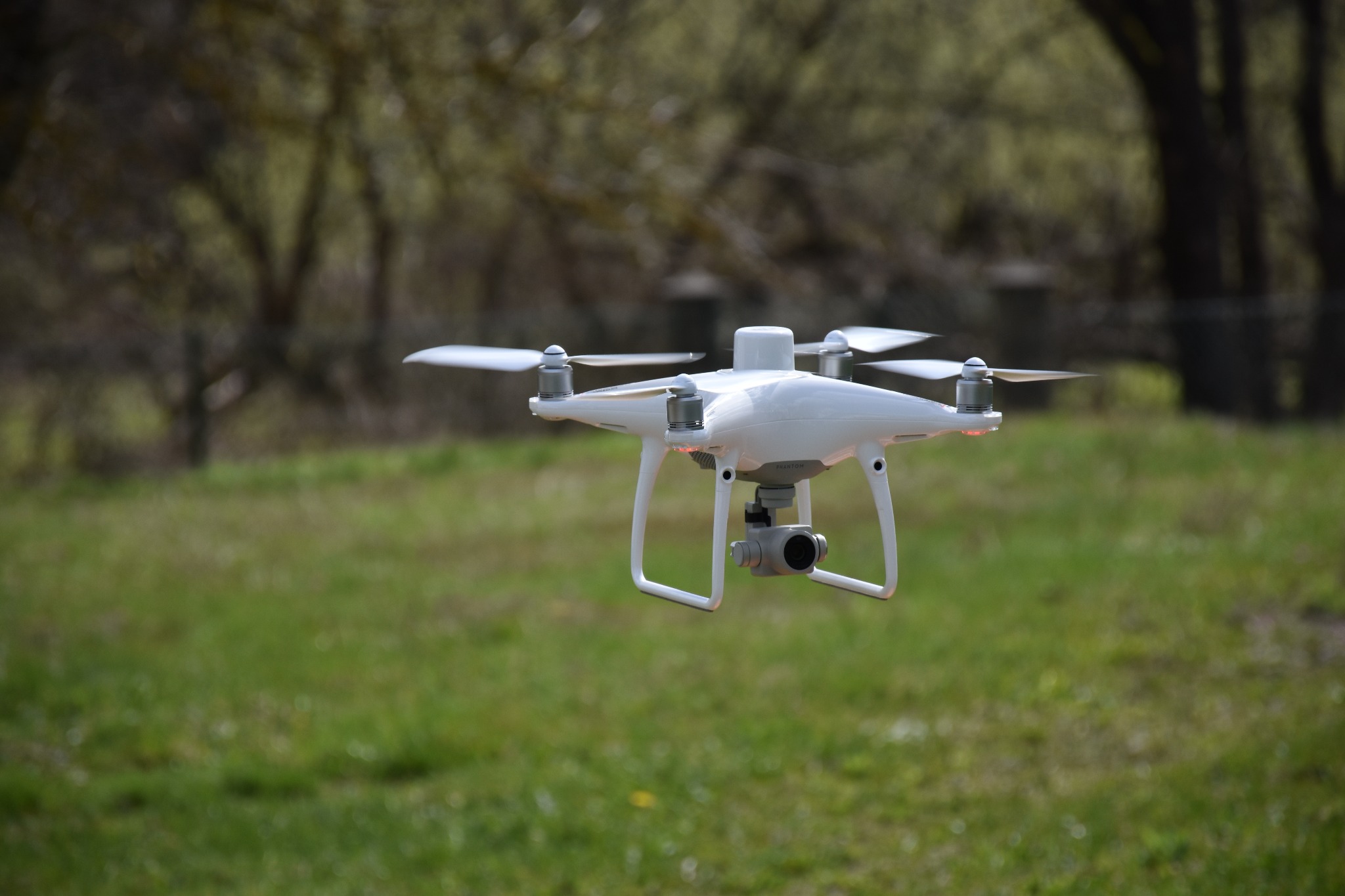 Biały dron z kamerą w locie nad zieloną trawą, w tle drzewa z ledwo rozwiniętymi liśćmi.